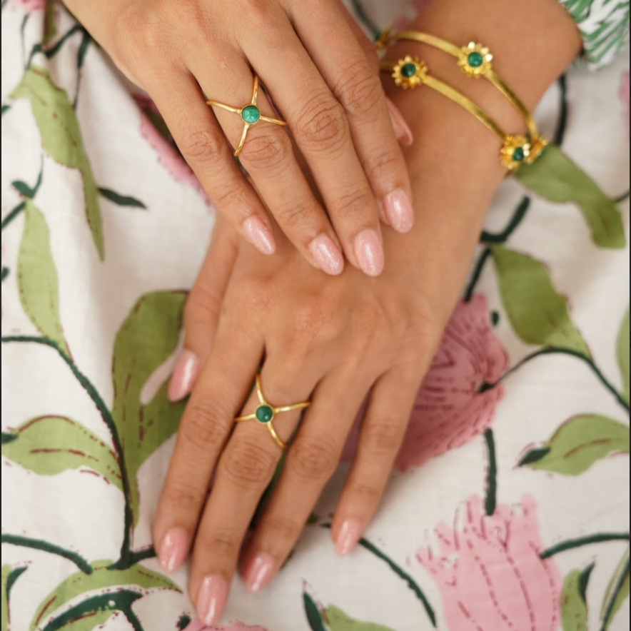 Close-up of hands wearing gold rings with green gemstones on a floral-patterned fabric background.